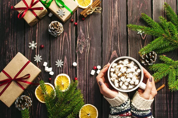Жіночі руки у вовняному светрі з чашкою гарячої кави з зефіром, різдвяні подарунки, ялинові гілки, конуси, апельсинові скибочки, сніжинки, сніг. Традиційне святкове прикраса Різдвяний темний — стокове фото
