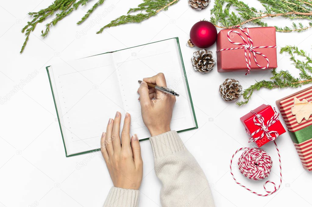 Female hands in knitted sweater are writing with pen in clean notebook plans for the new year, gift boxes, fir branches on white table flat lay. Christmas planning concept Holiday decorations 2019 Goals