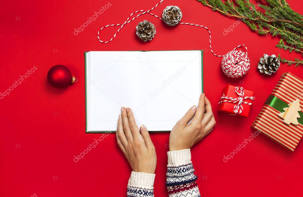 Female hands in knitted sweater holds open clean notebook, gift boxes, fir branches on red background flat lay Christmas planning concept Holiday decorations 2019 Goals winter