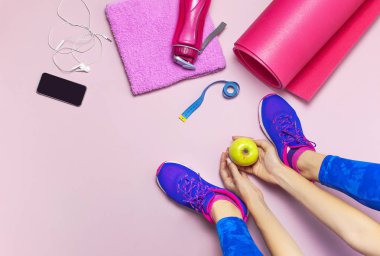 Genç kadın mavi spor ayakkabı içinde elma elinde, eğitim için hazırlanıyor tutar. Şişe su, yoga mat, telefon, pastel pembe arka plan düz kulaklıktan üstten görünüm yatıyordu. Spor ayakkabı Fitness kavramı