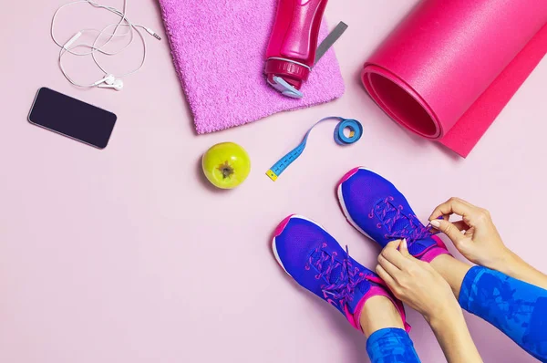 Genç kadın mavi spor ayakkabı içinde elma elinde, eğitim için hazırlanıyor tutar. Şişe su, yoga mat, telefon, pastel pembe arka plan düz kulaklıktan üstten görünüm yatıyordu. Spor ayakkabı Fitness kavramı