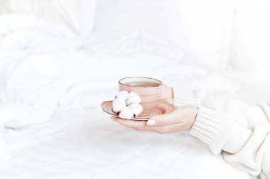 Beyaz örgü kazak kadınlar elinde fincan çay tabağı ile hassas pamuk çiçek yatakta üzerinde tutun. Rahat sabah kahvaltı evde yaşam tarzı nazik arka plan kopya alanı sonbahar kış kavramı
