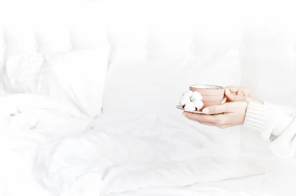 白いニットのセーターの女性手は ベッドで繊細なコットン花とソーサーに熱いお茶のカップを保持します 自宅の居心地の良い朝食ライフ スタイルに優しいバック グラウンド コピー スペース秋冬コンセプト — ストック写真