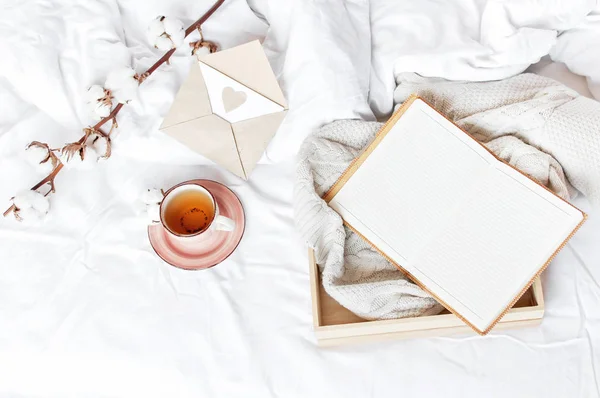 Cup of hot tea branch of cotton wooden tray knitted plaid sweater open notebook love letter in bed. Cozy morning breakfast at home Lifestyle gentle background Copy Space autumn winter concept