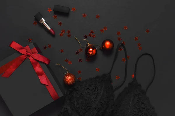 Black gift box with red ribbon, lace bra underwear, red lipstick, Christmas  balls, holographic confetti on dark background top view flat lay. Female  essential accessories, gift for new year christmas Stock Photo