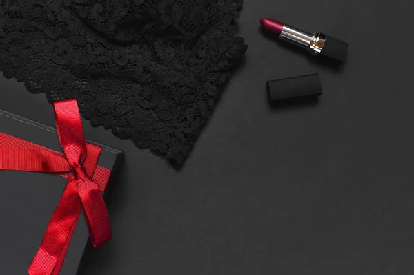 Black gift box with red ribbon, lace bra underwear, red lipstick, Christmas  balls, holographic confetti on dark background top view flat lay. Female  essential accessories, gift for new year christmas Stock Photo