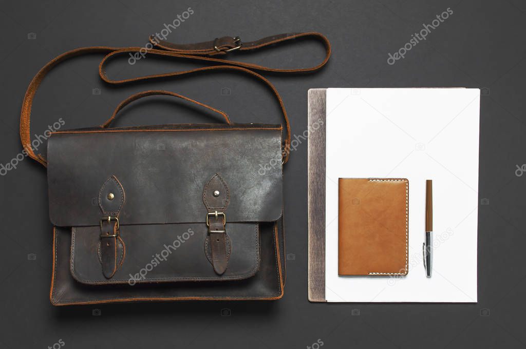 Fashionable concept. Brown leather men's bag, wristwatch, leather passport cover, pen, blank white sheets on black background top view flat lay with copy space Accessories businessman stylish clothes