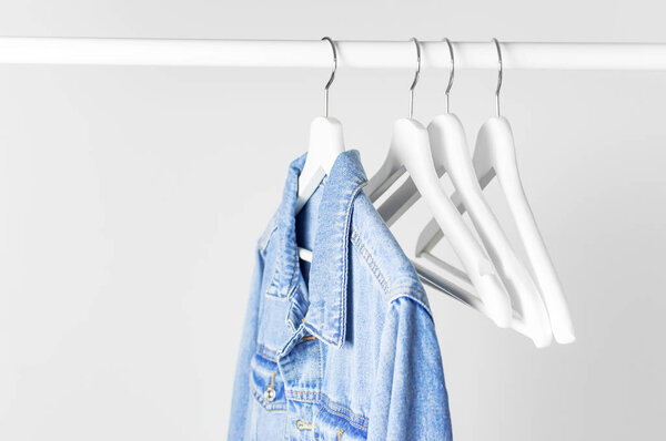 Blue denim jacket on white wooden coat hanger on a rod against light gray wall flat lay copy space. Denim, fashionable jacket, women's or men's trend clothing, fashion background. Store concept, sale