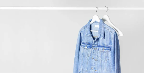 Blue denim jacket on white wooden coat hanger on a rod against light gray wall flat lay copy space. Denim, fashionable jacket, women's or men's trend clothing, fashion background. Store concept, sale