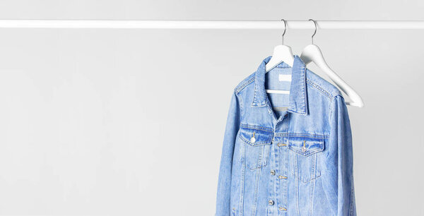 Blue denim jacket on white wooden coat hanger on a rod against light gray wall flat lay copy space. Denim, fashionable jacket, women's or men's trend clothing, fashion background. Store concept, sale