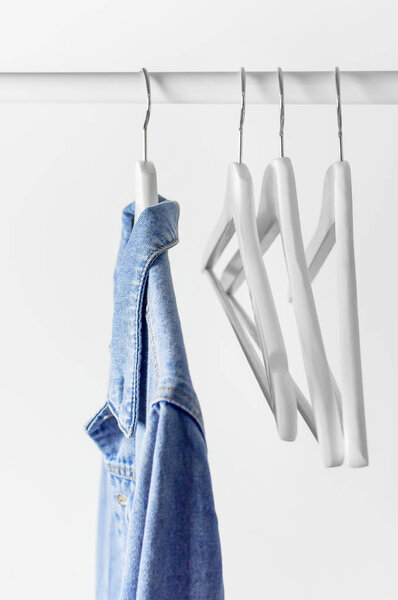 Blue denim jacket on white wooden coat hanger on a rod against light gray wall flat lay copy space. Denim, fashionable jacket, women's or men's trend clothing, fashion background. Store concept, sale