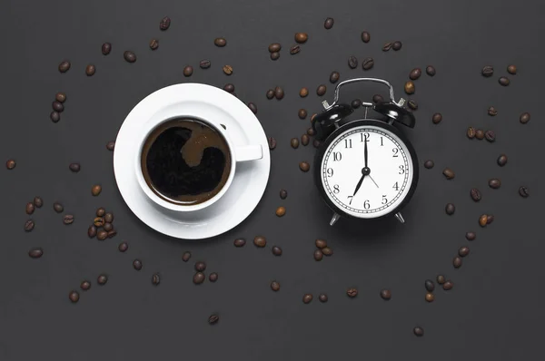 Flat lay cup of black coffee, coffee beans, black alarm clock on gray dark background top view copy space. Minimalistic food concept, morning breakfast, time to work, hot drink coffee background