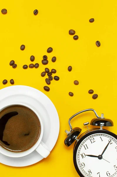 Flat lay cup of black coffee, coffee beans, black alarm clock on yellow background top view copy space. Minimalistic food concept, morning breakfast, time to work, hot drink, coffee background.
