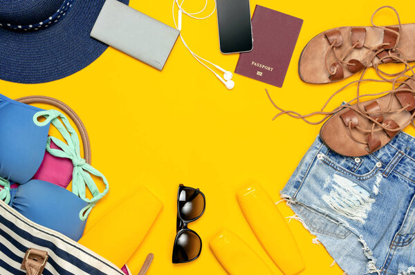 Female summer clothing accessories on yellow background. Concept of travel vacation sea. Hat swimming suit denim shorts leather sandals sunglasses sunscreen sea bag passport phone. Flat lay top view