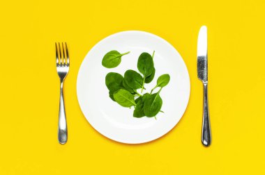 Beyaz çatal bıçaklı taze yeşil ıspanak yaprakları parlak sarı arka plan düz görünüm fotokopi alanında yatıyordu. Yavru ıspanak yaprakları, sağlıklı yiyecekler, diyet. Yaratıcı gıda konsepti.