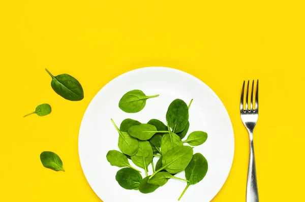 Fresh green spinach leaves on a white plate fork on bright yellow background flat lay top view copy space. Baby young spinach leaves, healthy food, diet. Creative food concept.