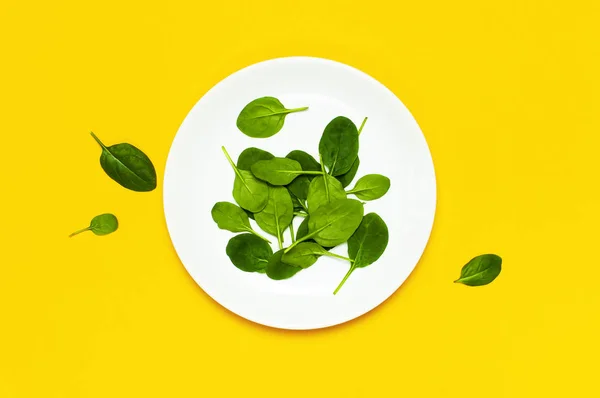 Fresh green spinach leaves on white plate on bright yellow background flat lay top view copy space. Baby young spinach leaves, Ingredient for salad, healthy food, diet. Creative food concept.