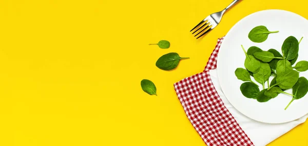 Fresh green spinach leaves on a white plate fork kitchen towel on bright yellow background flat lay top view copy space. Baby young spinach leaves, healthy food, diet. Creative food concept.