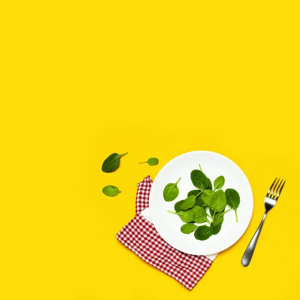 Fresh green spinach leaves on a white plate fork kitchen towel on bright yellow background flat lay top view copy space. Baby young spinach leaves, healthy food, diet. Creative food concept.