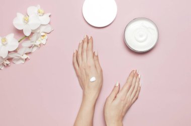 Genç kadın elini nemlendirir ile kozmetik krem losyon krem vücut süt beyaz Phalaenopsis orkide çiçek pembe arka plan düz üzerinde açılan kapsayıcıyla üstten görünüm minimalizm tarzı güzellik kavramı yatıyordu.