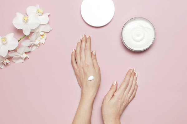 Young woman moisturizes her hand with cosmetic cream lotion opened container with cream body milk White Phalaenopsis orchid flowers on pink background Flat lay top view minimalism style Beauty concept