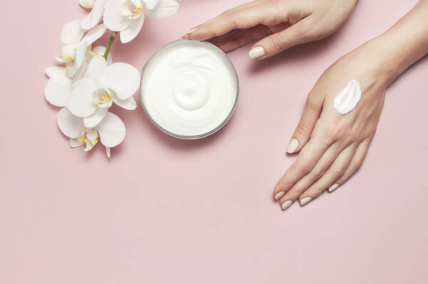 Young woman moisturizes her hand with cosmetic cream lotion opened container with cream body milk White Phalaenopsis orchid flowers on pink background Flat lay top view minimalism style Beauty concept