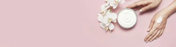 Mujer joven hidrata su mano con crema cosmética recipiente abierto con crema cuerpo leche blanco Phalaenopsis flores de orquídea sobre fondo rosa plano vista superior minimalismo estilo concepto de belleza — Foto de Stock