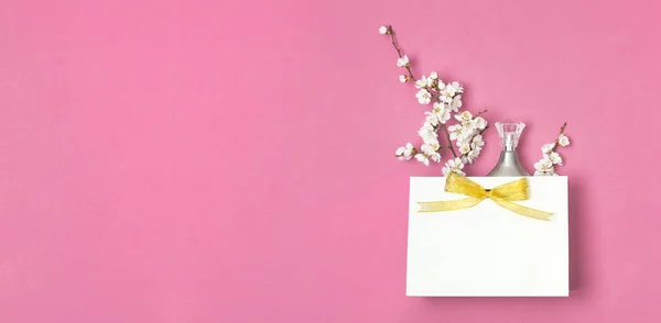 Weiße Geschenktüte mit Goldband, eine Flasche Parfüm, weiße Frühlingsblumen auf leuchtend rosa Hintergrund. Grußkarte mit zarten Blumen rosa floralen Hintergrund. Frühling minimales Konzept flache Lageansicht von oben — Stockfoto