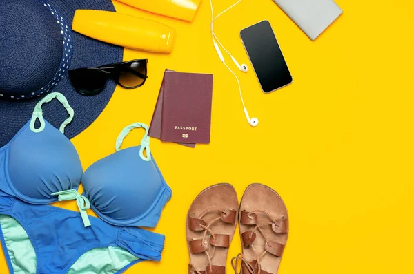 Sarı arka planda kadın yaz plaj aksesuarları. Seyahat tatil deniz kavramı. Mavi şapka yüzme takım elbise deri sandalet güneş gözlüğü güneş koruyucu pasaport telefon kulaklıklar düz yatıyordu üst görünüm