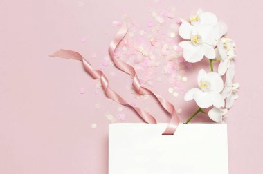 Düz üst görünüm Beyaz Phalaenopsis orkide çiçek pastel pembe arka plan üzerinde şenlikli konfeti beyaz hediye çantası yatıyordu. Orkide Tropikal çiçek ile tebrik kartı. Tatil doğum günü Kadınlar Günü çiçek Kartı