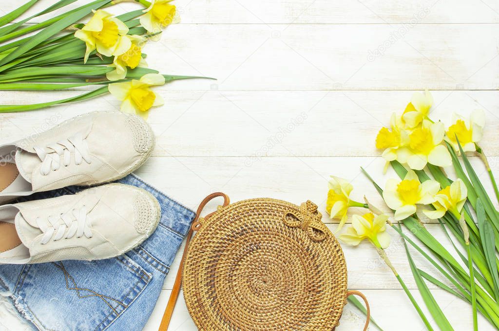 Fashionable natural organic round rattan bag, denim shorts, beige women's espadrilles, yellow narcissus daffodil flowers on light wooden background flat lay. Summer fashion concept. Ecobags from Bali