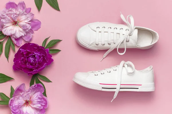 White female fashion sneakers and pink purple flowers peonies on pink background. Flat lay, top view, copy space. Women's shoes. Stylish white sneakers. Fashion blog or magazine concept.