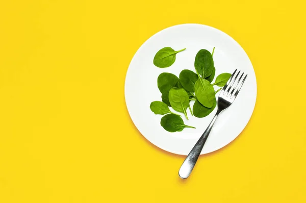 Verse groene spinazie bladeren op een witte plaat vork op heldere gele achtergrond plat leggen bovenaanzicht kopie ruimte. Baby jonge spinazie bladeren, gezonde voeding, dieet. Creatieve voedsel concept. — Stockfoto