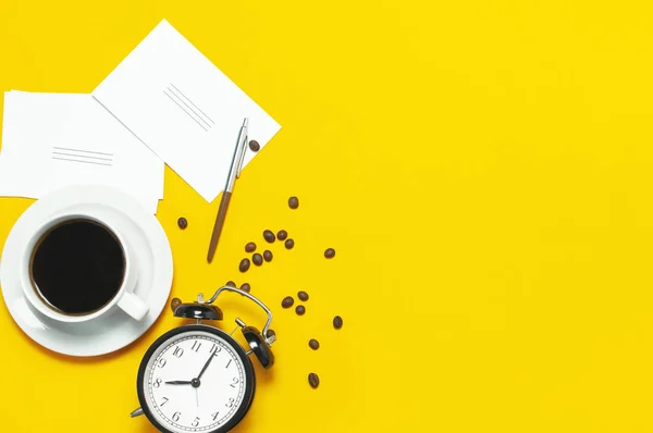 Flat lay taza de café negro, granos de café, reloj despertador negro, bolígrafos, tarjetas blancas sobre fondo amarillo vista superior espacio de copia. Concepto creativo tiempo para trabajar, escritorio femenino, fondo de café — Foto de Stock