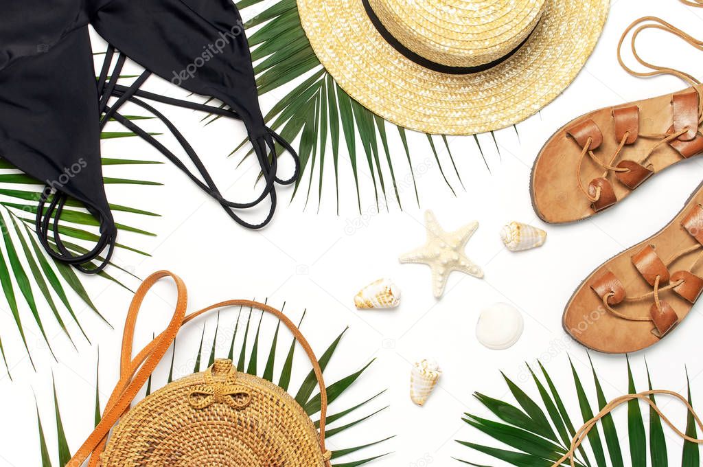 Woman's beach accessories flat lay. Round trendy rattan bag straw hat black swimsuit leather sandals tropical palm leaves seashells on white background. Top view copy space. Summer backdrop