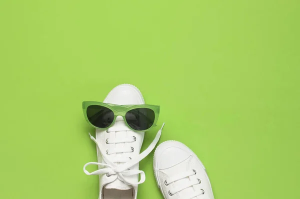 Witte vrouwelijke mode sneakers en trendy zonnebrillen op een groene achtergrond. Platte lay Top View Kopieer ruimte. Damesschoenen, accessoires. Stijlvolle witte sneakers. Creatief concept mode blog of tijdschrift — Stockfoto