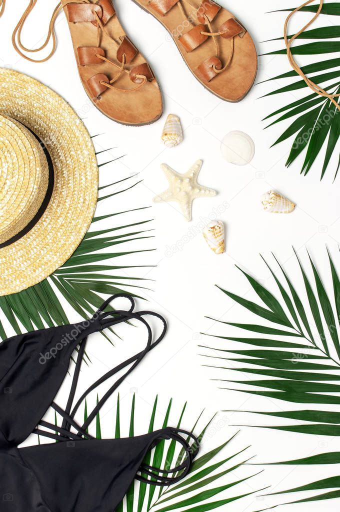 Woman's beach accessories flat lay. Round trendy rattan bag straw hat black swimsuit leather sandals tropical palm leaves seashells on white background. Top view copy space. Summer backdrop