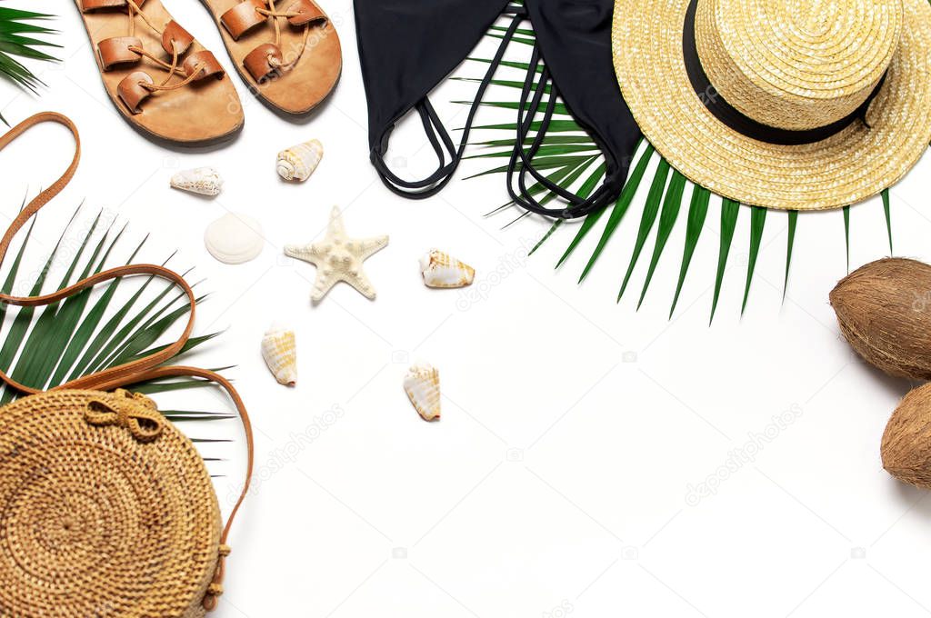Woman's beach accessories flat lay. Round trendy rattan bag straw hat black swimsuit leather sandals tropical palm leaves coconut seashells on white background. Top view copy space. Summer backdrop