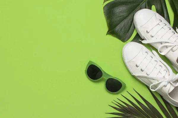 Beyaz kadın moda spor ayakkabıları, trendy güneş gözlüğü, yeşil arka plan üzerinde tropikal yaprakları. Düz lay üst görünüm kopyalama alanı. Kadın ayakkabıları, aksesuarlar. Şık beyaz spor ayakkabılar. Yaratıcı yaz konsepti — Stok fotoğraf