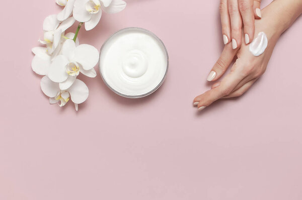 Young woman moisturizes her hand with cosmetic cream lotion opened container with cream body milk White Phalaenopsis orchid flowers on pink background Flat lay top view minimalism style Beauty concept
