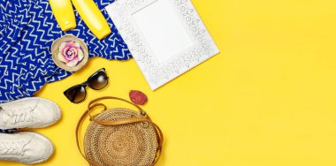 Kadın yaz moda arka plan. Sarı arka planda giysi ve aksesuarlar. Mavi elbise, yuvarlak rattan çanta, espadrilles ayakkabı, güneş gözlüğü, güneş kremi, ahşap resim çerçevesi. Düz lay üst görünüm