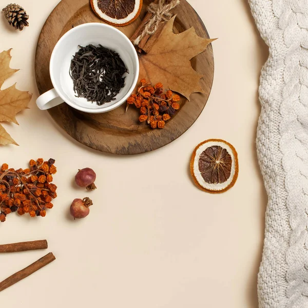 Composition plate de l'automne. Tasse blanche avec feuilles de thé, assiette en bois, pull ou plaid tricoté, feuilles d'érable jaune, cannelle, frêne sec de montagne, orange sur fond beige — Photo