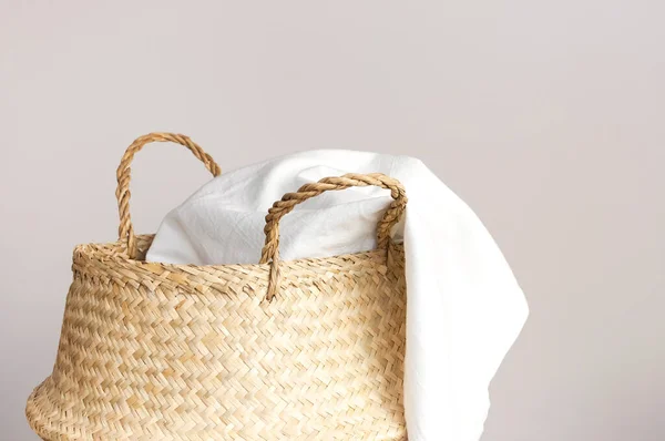 Straw wicker basket and white natural cotton fabric, towel on gray background. Fashionable bamboo basket stylish interior item eco design handmade. Decor of home. Natural eco materials, storage basket.