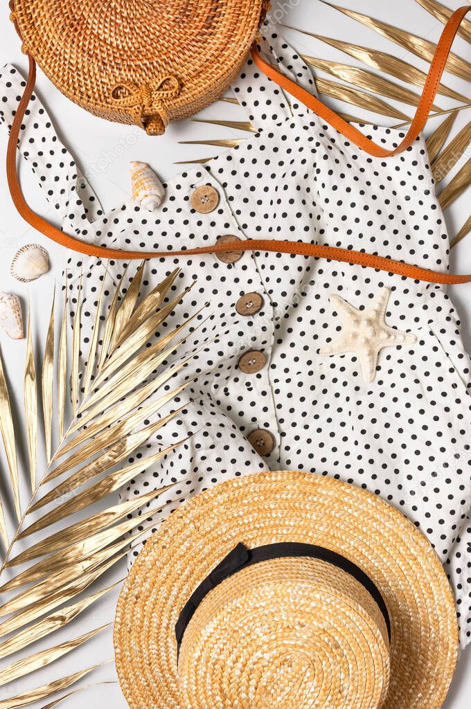 Summer women's white dress in black peas rattan woven bag brown sandals straw hat golden palm leaf shells starfish on light background. Flat lay top view. Women's beach fashion, travel vacation.