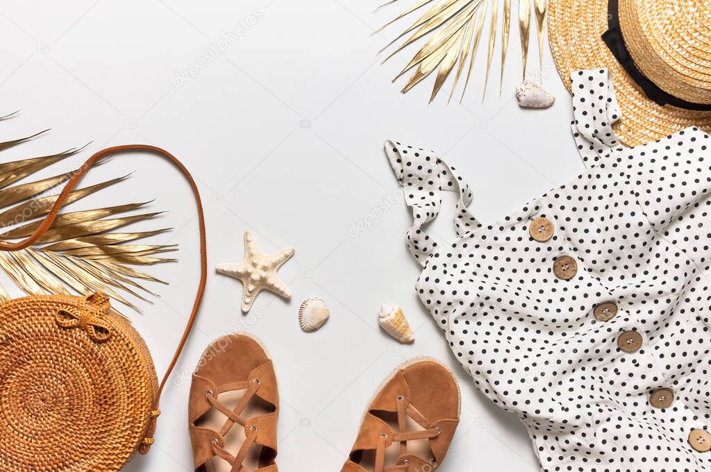 Summer women's white dress in black peas rattan woven bag brown sandals straw hat golden palm leaf shells starfish on light background. Flat lay top view. Women's beach fashion, travel vacation.