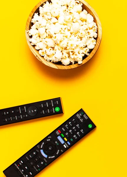Black TV, audio remote control and popcorn on bright yellow background flat lay top view copy space. Minimalistic background with a remote control, watching a movie, series set-top boxes audio system.