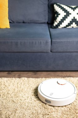 Beyaz robotik elektrikli süpürge oturma odasını gri kanepe ve rahat halının zeminine karşı temizler. Ev Temizliği Konsepti. Yeni teknolojiler, hızlı ev temizleme otomatik robot.