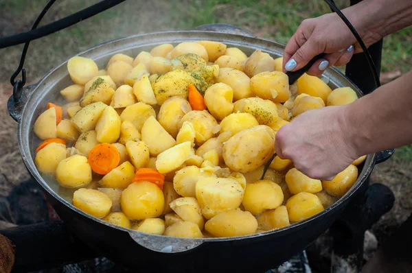 Camping kitchen. Cooking potatoes with carrots in a large metal pot on the nature. Hands with a spoon.