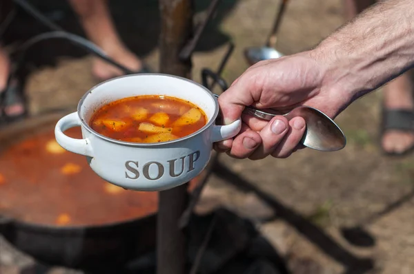 Traditional red soup in a large metal cauldron on a bonfire. Cooking on a camping trip. Hand holds a plate in which soup is poured. The text on the bowl - soup.