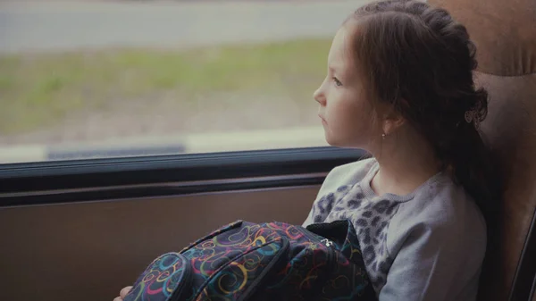 Jonge, mooie meisje passagier met schooltas in de bewegende schoolbus kijkt uit het raam — Stockfoto